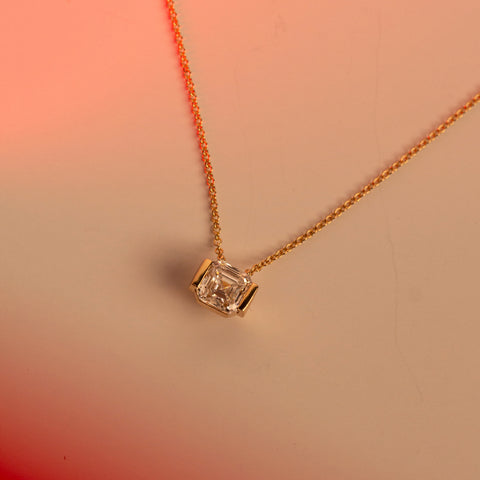 A delicate gold chain necklace with a single asscher-cut lab-grown diamond pendant in a gold half-bezel setting, displayed on a warm peach and pink gradient background.