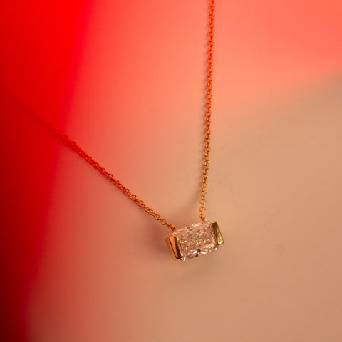 A delicate gold chain necklace with a rectangular radiant-cut lab-grown diamond pendant, shown against a warm beige background with a red light gradient on the left.