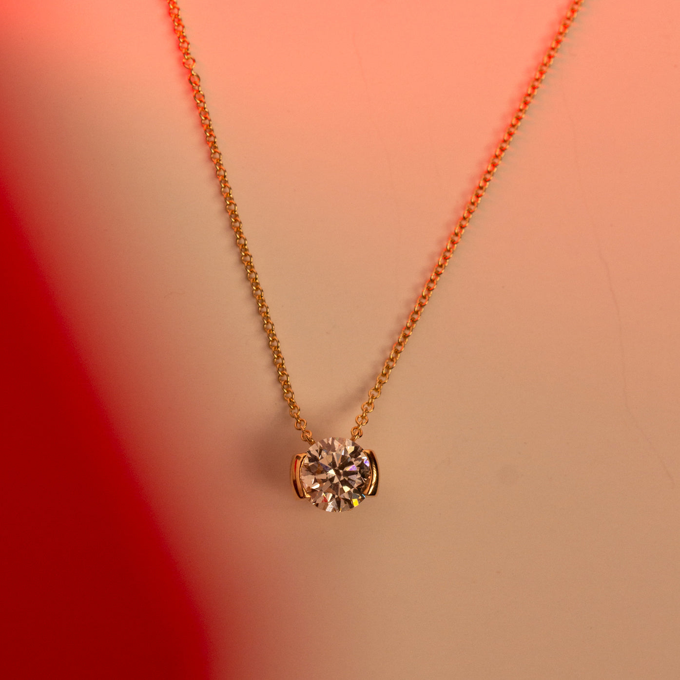 A delicate gold chain necklace with a single round lab grown diamond pendant, displayed against a warm peach and red gradient background.