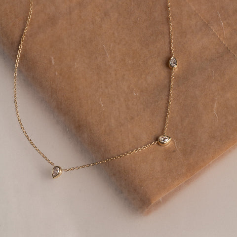 A delicate gold chain necklace with three small pear-cut lab-grown clear diamonds, draped over a brown textured fabric surface.